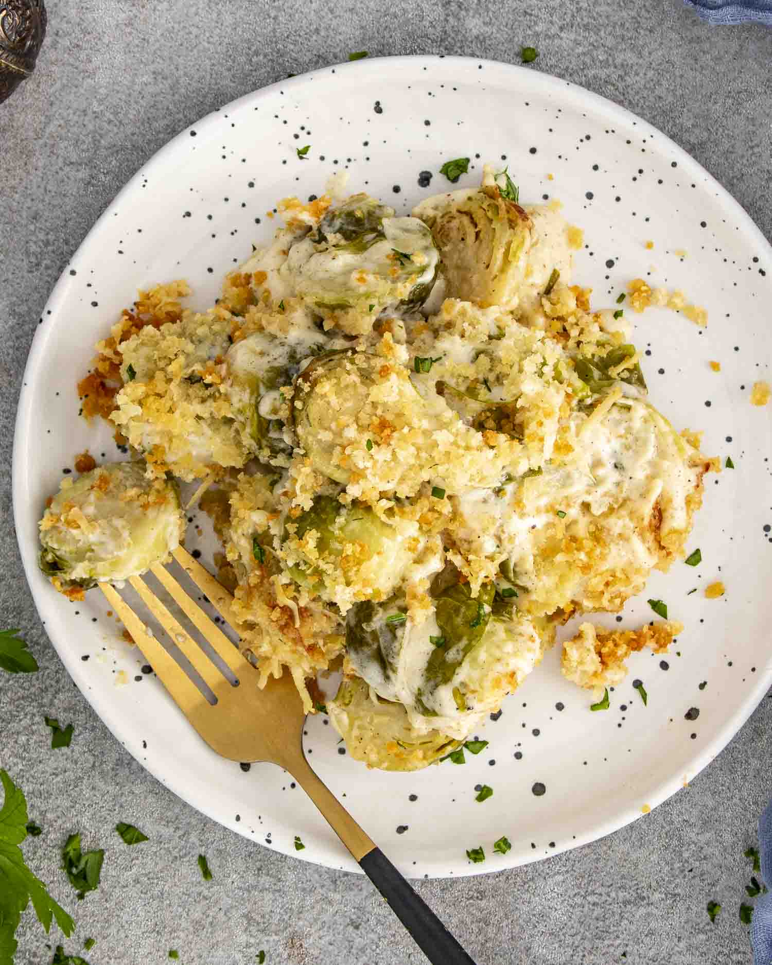 Plate of creamy Brussels sprouts gratin topped with Gruyère, Parmesan, and crispy breadcrumbs, served with gold fork and parsley garnish.