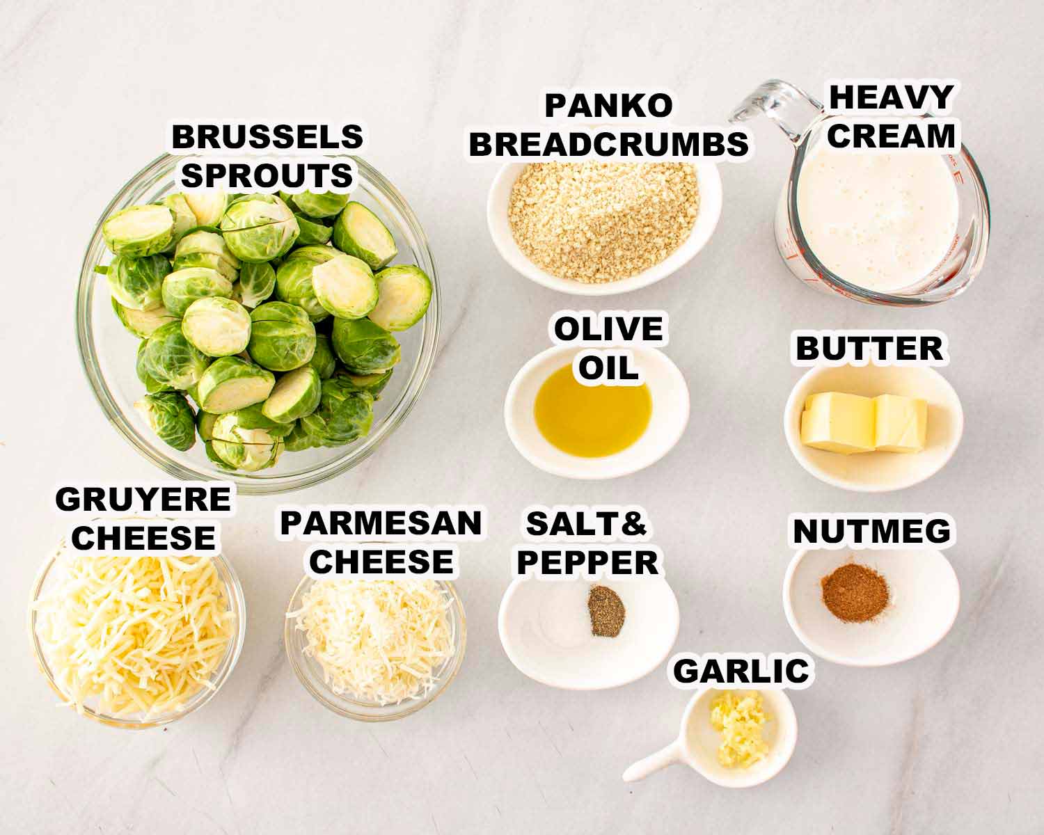 Overhead view of Brussels sprouts gratin ingredients including Brussels sprouts, Gruy&egrave;re, Parmesan, cream, garlic, butter, and breadcrumbs on white surface.