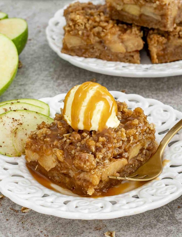 Slice of caramel apple crisp bar topped with vanilla ice cream and caramel drizzle, served with fresh apple slices.