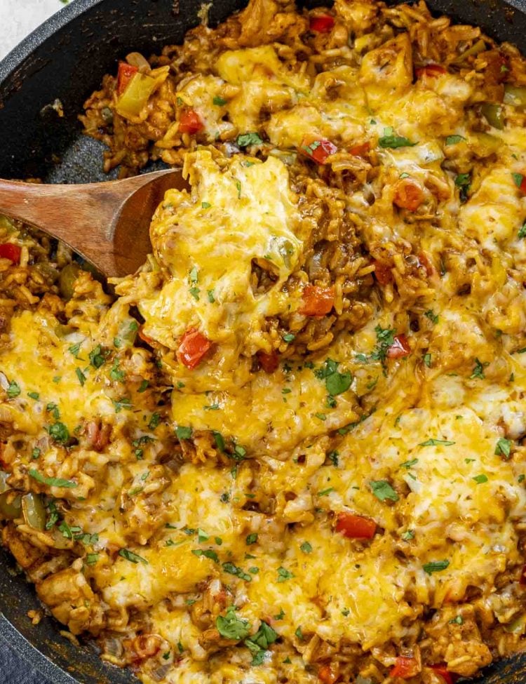 A skillet filled with Chicken Fajita Rice Skillet, featuring vibrant red and green bell peppers, melted cheddar cheese, tender chicken, and fluffy rice garnished with fresh cilantro. A wooden spoon is scooping up a cheesy, hearty portion of the dish.