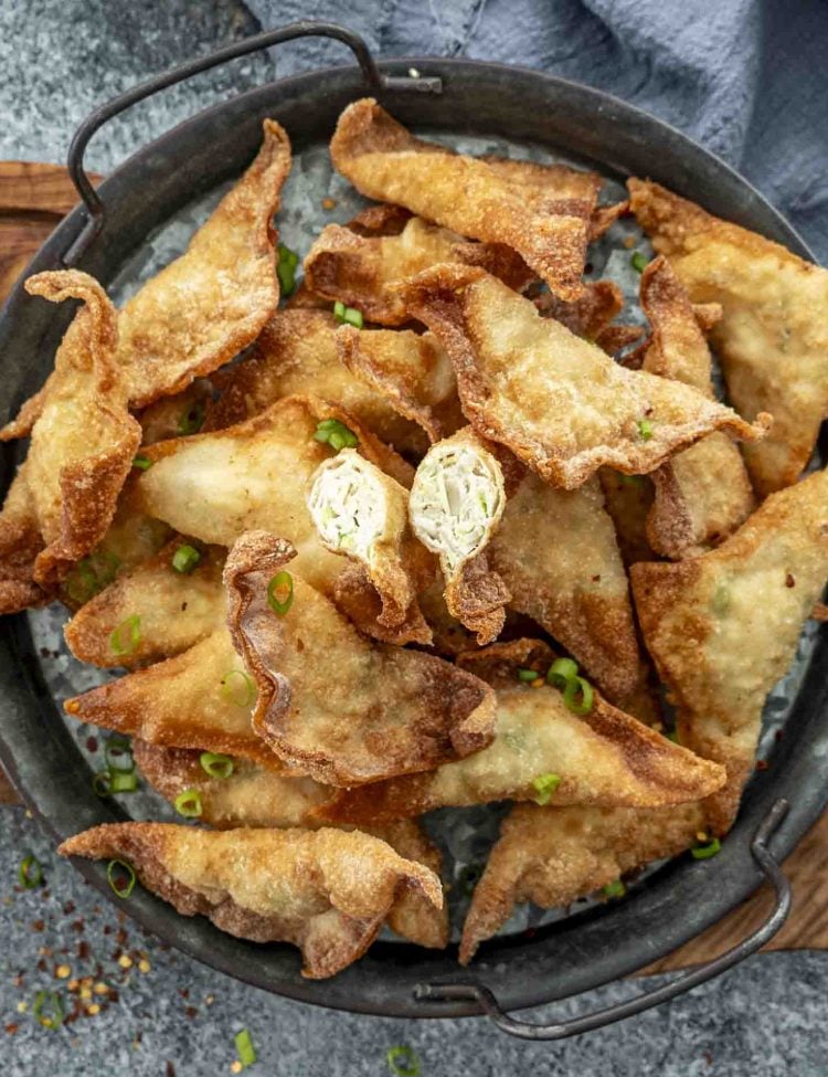 A black metal serving tray filled with crispy, golden-brown chicken fried wontons, garnished with finely chopped green onions. One wonton is cut open, revealing a juicy, well-seasoned chicken and cabbage filling. A small bowl of dipping sauce with chili flakes sits in the background, adding a rich, savory contrast to the crunchy wontons.