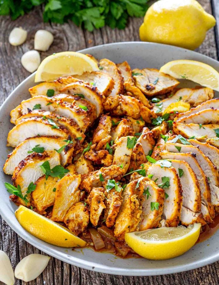 sliced chicken shawarma in a white bowl garnished with parsley and lemon wedges.