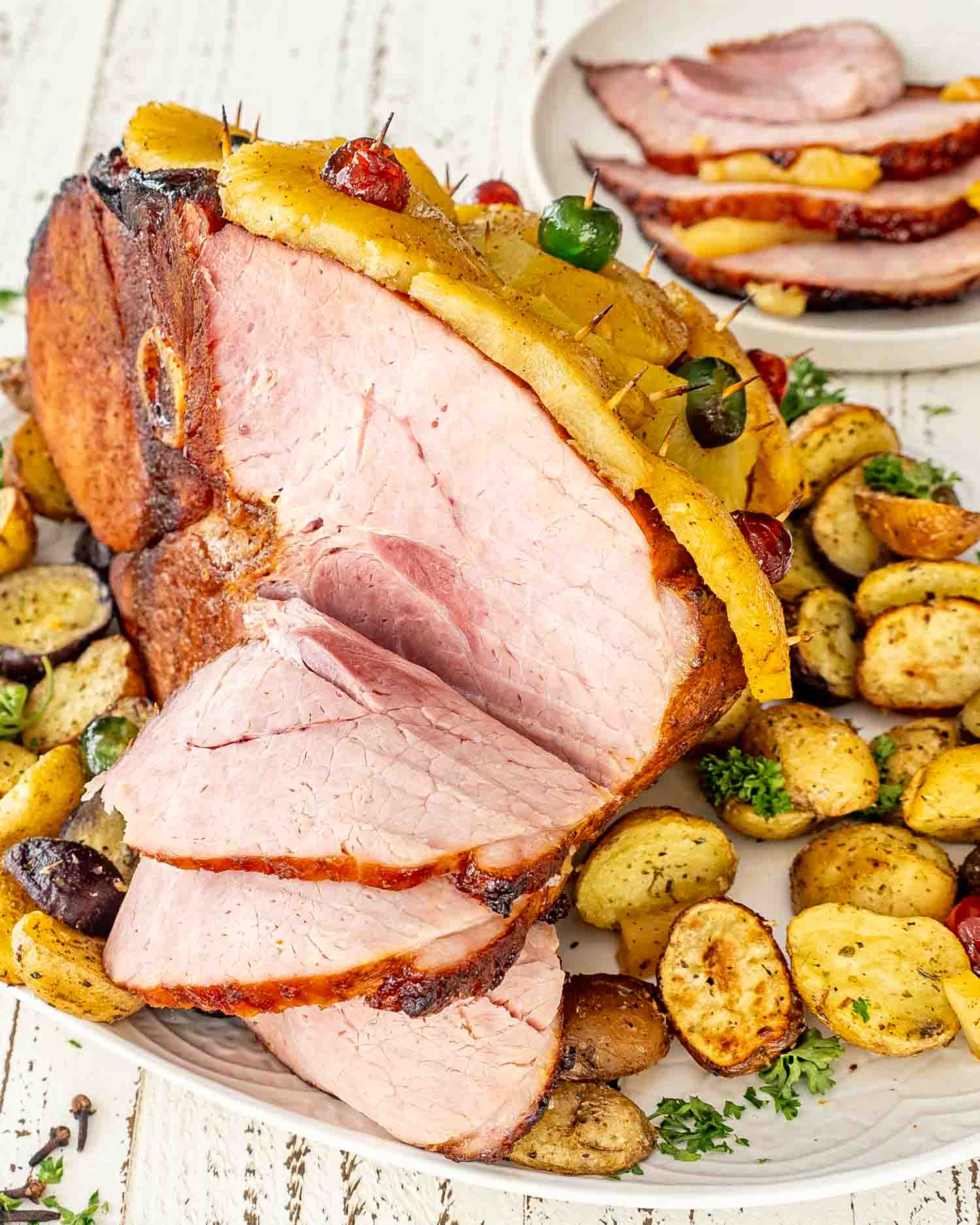 a whole christmas ham, some of it sliced along some roasted potatoes on a white platter.