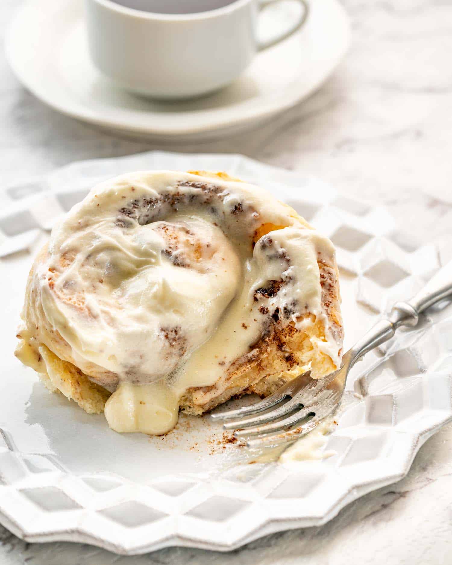 a cinnabon cinnamon roll on a white dessert plate with a cup of coffee in the background.