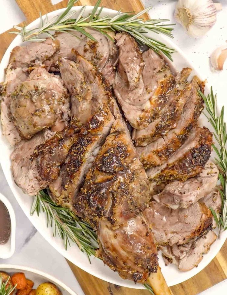 Sliced roasted leg of lamb on a white platter, garnished with rosemary sprigs, garlic cloves, and served with pan gravy.