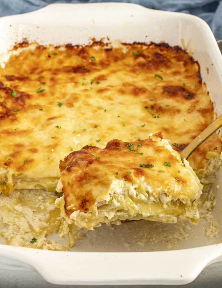 Creamy Gruy&egrave;re and leek scalloped potatoes being lifted from the baking dish, showing tender layers and browned cheesy top.