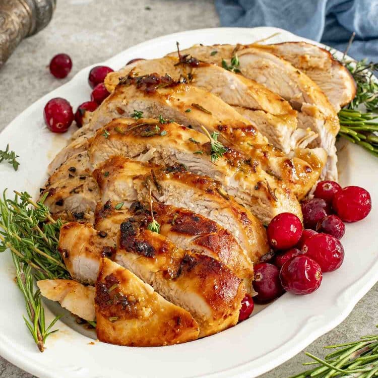 Juicy slices of Maple Dijon Glazed Turkey Breast arranged on a platter with rosemary, thyme, and bright red cranberries.