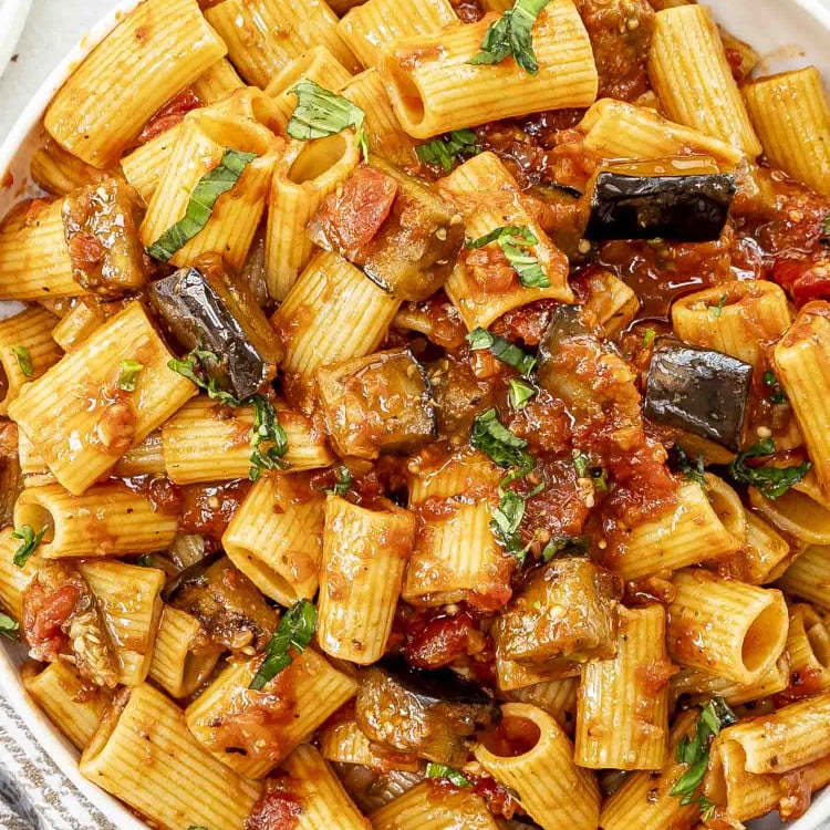 A white bowl of Pasta alla Norma featuring rigatoni in a rich tomato sauce with tender eggplant chunks, garnished with fresh basil.