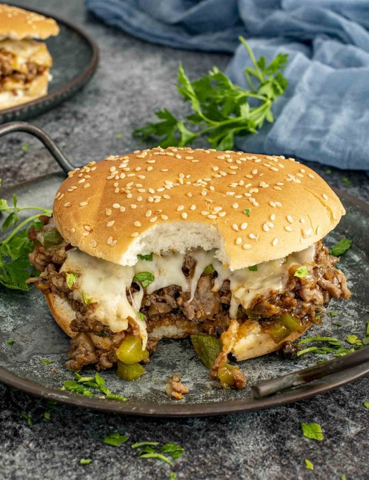 Philly Cheesesteak Sloppy Joe with a bite taken out, showing the cheesy, saucy beef filling with peppers and onions on a soft sesame bun.