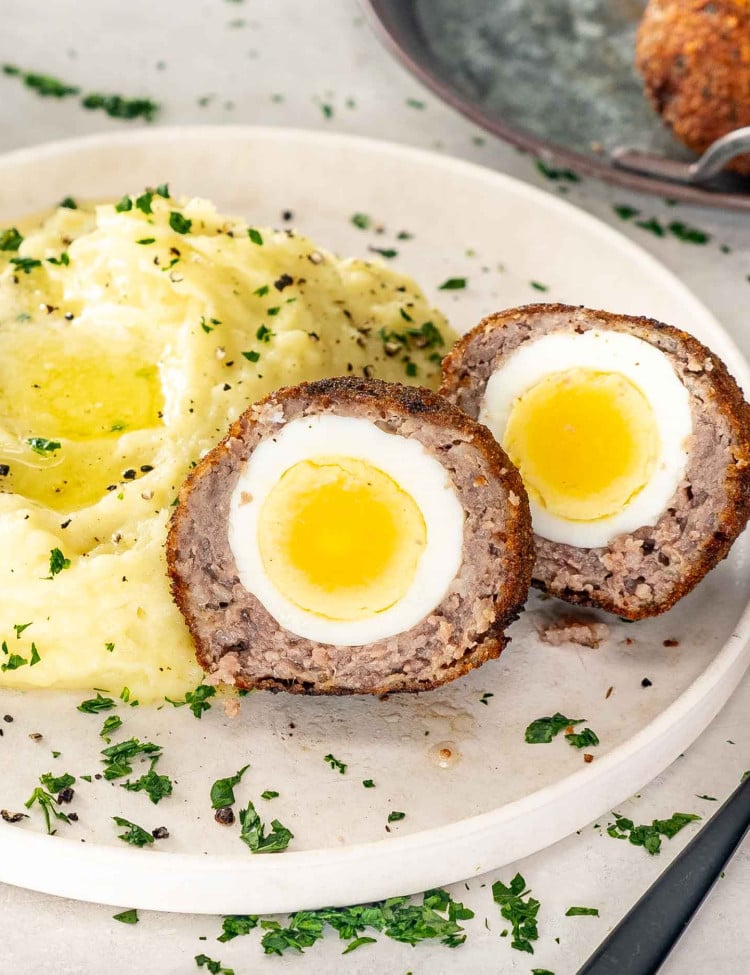a scotch egg cut in half on a white plate along side some mashed potatoes garnished with parsley.