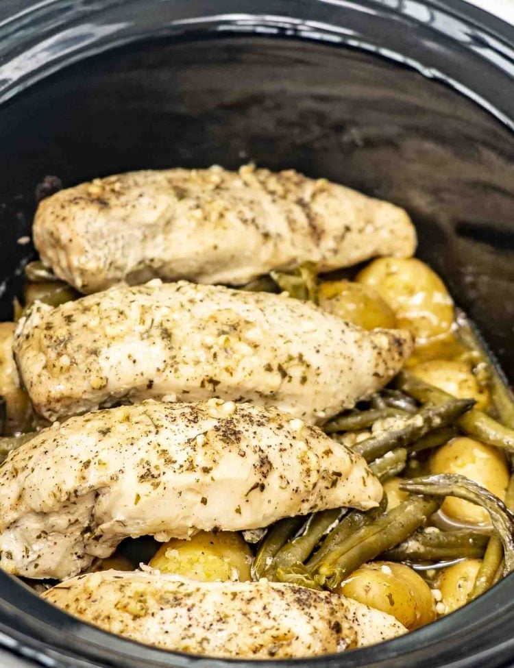 Close-up of slow-cooked chicken breasts, golden baby potatoes, and green beans, all seasoned with garlic, ranch, and butter, inside a black slow cooker.