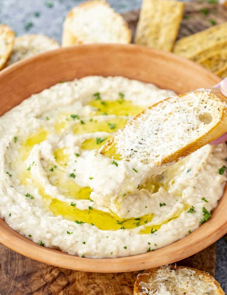 Bread slice dipped into creamy white bean dip, coated generously, with grated cheese and olive oil swirl.