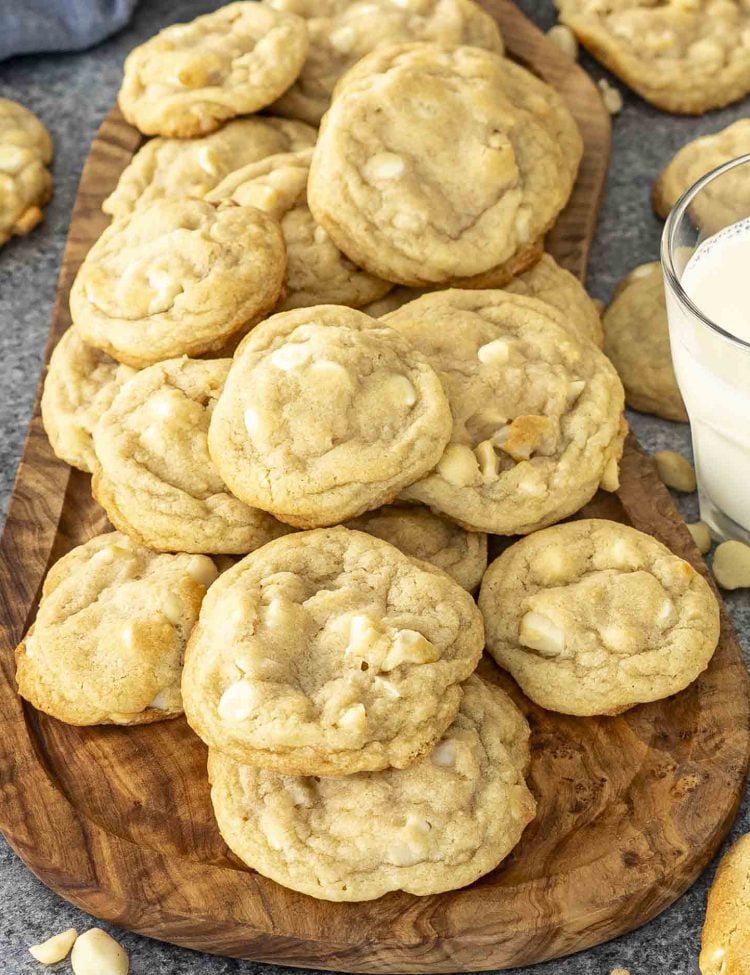 A wooden board piled high with freshly baked white chocolate macadamia nut cookies beside a glass of milk.